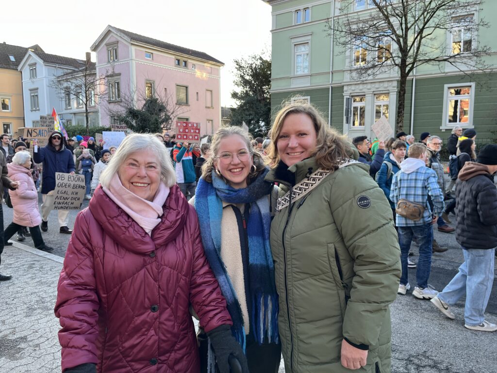Drei Frauen schauen auf einer Demo lächelnd in die Kamera.